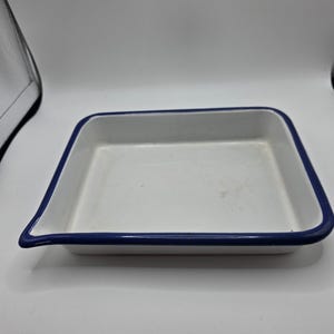 May include: A rectangular white baking dish with a smooth interior and a thick, dark blue rim. The dish features a small, pointed pouring spout on the left side, providing a functional design element for transferring liquids or sauces easily.
