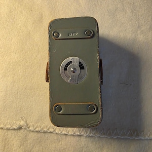 May include: A rectangular gray leather camera case featuring a circular silver exposure calculator dial on the front surface. The case includes two horizontal gray leather straps secured with metal rivets and the word JAPAN embossed on the top strap.