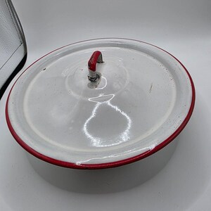 May include: A round white enamel pot with a matching lid. The lid features a red handle and a red rim. The pot has a smooth, glossy finish with visible light reflections on the surface. The design is simple, functional, and circular in shape.