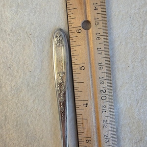 May include: A silver-toned metal spoon handle featuring an ornate, embossed floral and geometric pattern. The handle is positioned alongside a wooden ruler, showing a length of approximately 5.5 inches from the top to the start of the bowl.
