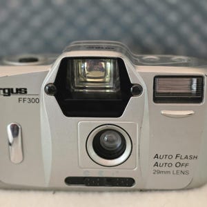 May include: A silver Argus FF300 compact film camera featuring a 29mm lens, an integrated flash unit, and a central viewfinder. The front casing displays the brand name Argus, the model FF300, and the text Auto Flash Auto Off 29mm Lens.