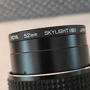 May include: A black camera lens filter with white text that reads HOYA 52mm SKYLIGHT (1B) JAPAN. The filter has a textured grip ring on the lower section and a smooth metal rim on the upper section where the product information is printed.