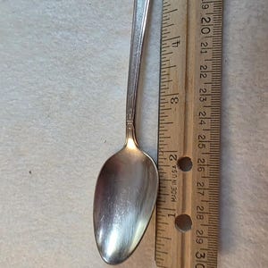 May include: A silver-toned metal spoon with an ornate, textured handle design. The spoon is positioned alongside a wooden ruler, showing a total length of approximately 5.75 inches from the tip of the bowl to the end of the handle.