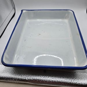 May include: A rectangular enamel baking pan with a glossy white interior and a deep blue rim. The surface of the pan features a smooth finish with minor signs of wear, including small dark specks scattered across the bottom of the basin.