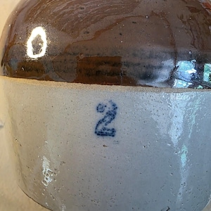 May include: A stoneware crock featuring a two-tone design with a glossy dark brown glazed upper section and a light grey glazed lower body. The lower portion displays a stamped blue number 2. The vessel has a rounded shape and a smooth texture.