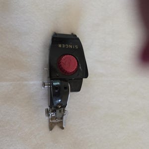 May include: A black Singer sewing machine presser foot with a red knob. The word "SINGER" is printed in white on the top of the foot. The foot has a silver metal base and is designed for sewing.