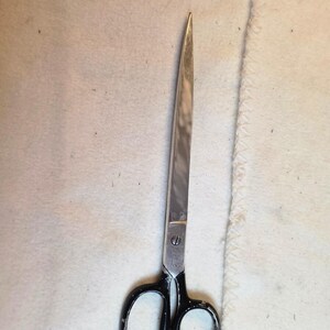 May include: A pair of metal sewing scissors with long, pointed silver blades and black handles. The scissors feature a central screw pivot point and a small, faint inscription on the metal near the handle, showing signs of wear on the black finish.