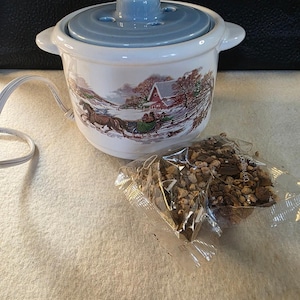 May include: A white ceramic electric potpourri warmer with a light blue lid and a winter scene featuring a horse-drawn sleigh and a red barn. A clear plastic packet containing dried cinnamon and spice potpourri sits in front of the warmer.