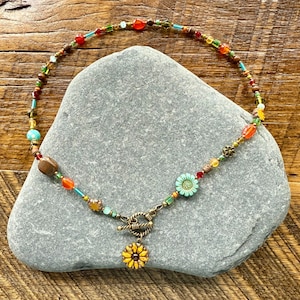 May include: A vibrant beaded necklace with a sunflower pendant. The necklace incorporates a mix of colourful beads in red, orange, yellow, green, and blue. The pendant is a small, detailed sunflower with yellow petals and a brown centre.