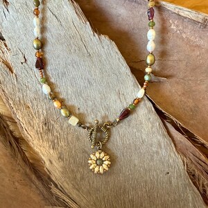 Peut inclure: Un collier de perles avec un mélange de perles blanches, vertes et marron, avec un pendentif tournesol doré. Le collier a un fermoir à bascule et est présenté sur une surface en bois.