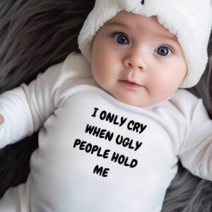 Op de afbeelding: Een baby in een witte romper met de tekst "I ONLY CRY WHEN UGLY PEOPLE HOLD ME". De baby draagt ook een witte berenmuts met zwarte accenten. De baby ligt op een grijze, harige ondergrond.