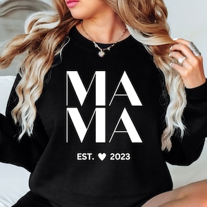 Op de afbeelding: Zwart sweatshirt met het woord "MAMA" in grote witte letters. Daaronder staat de tekst "EST. ♥ 2023". De sweatshirt heeft een ronde hals.