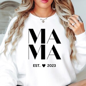 Op de afbeelding: Wit sweatshirt met zwarte tekst die "MAMA EST. 2023" luidt met een hartje tussen de woorden "EST." en "2023".