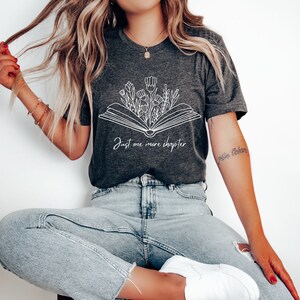 Może przedstawiać: Szary t-shirt z białym nadrukiem otwartego książki z kwiatami wyrastającymi z niej. Pod nadrukiem znajduje się tekst "Just one more chapter".