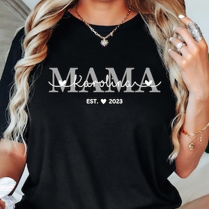 May include: Black t-shirt with white text that reads "MAMA Karolina EST. 2023" with a heart symbol between EST and 2023.