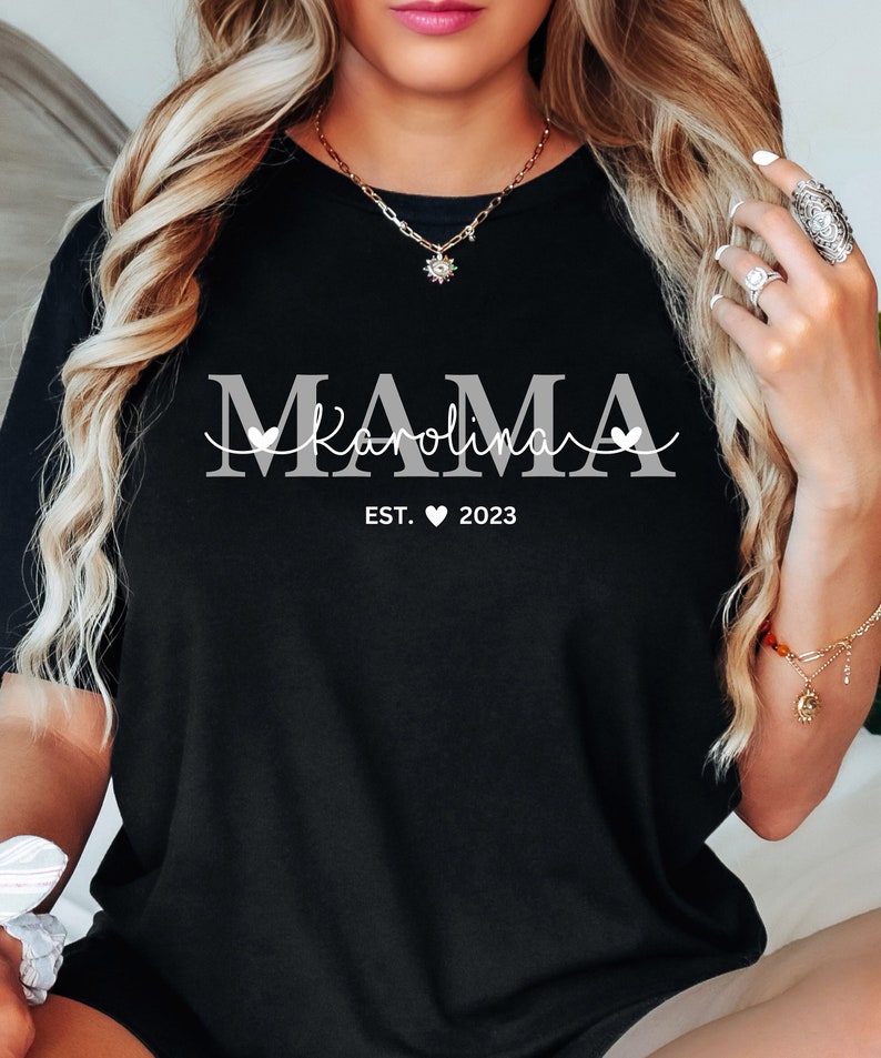 May include: Black t-shirt with white text that reads "MAMA Karolina EST. 2023" with a heart symbol between EST and 2023.