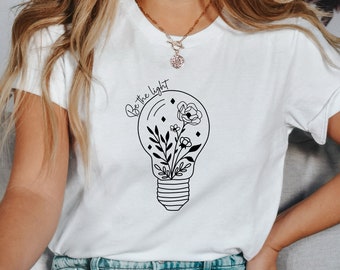 May include: White t-shirt with a black graphic of a light bulb with flowers and leaves inside. The text "Be the light" is written in a cursive font around the light bulb.