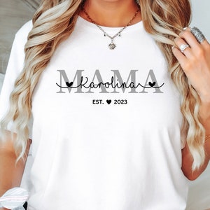 May include: White t-shirt with the text "MAMA Karolina EST. 2023" in gray and black lettering. The text includes a heart symbol.