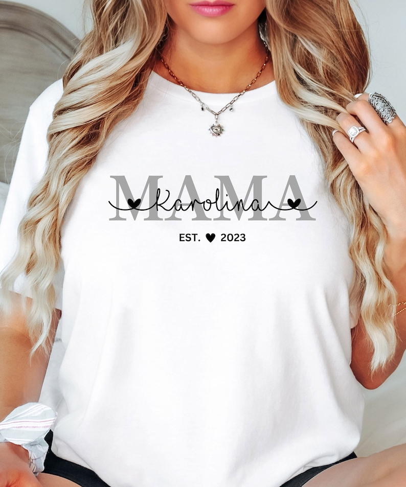 May include: White t-shirt with the text "MAMA Karolina EST. 2023" in gray and black lettering. The text includes a heart symbol.