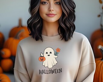May include: A woman wearing a beige sweatshirt with a cartoon ghost holding a pumpkin and a flower. The sweatshirt has the word "HALLOWEEN" printed on it. She is standing in front of a wall with pumpkins.