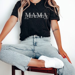 May include: A black t-shirt with the text "MAMA" in white with the dates "EST. 2020" and "EST. 2023" on either side of the word "MAMA".