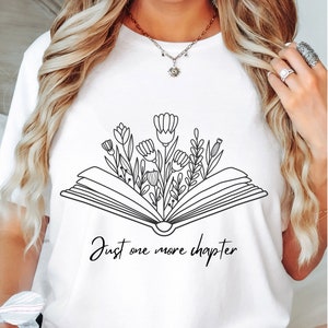 Może przedstawiać: Biała koszulka z czarnym rysunkiem otwartego książki, z kt&oacute;rej wyrastają kwiaty. Pod książką widnieje napis "Just one more chapter".