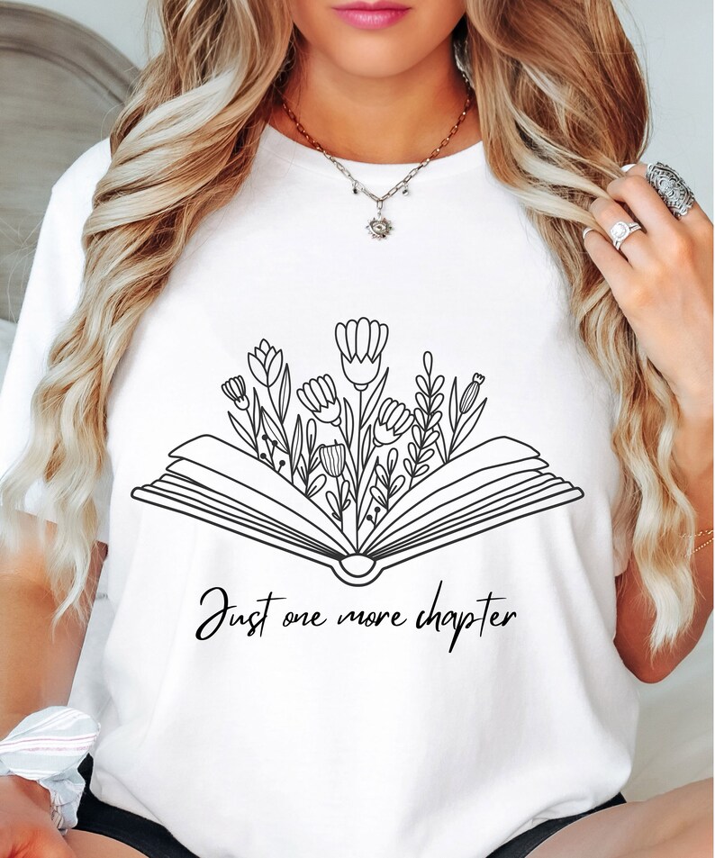 Może przedstawiać: Biała koszulka z czarnym rysunkiem otwartego książki, z kt&oacute;rej wyrastają kwiaty. Pod książką widnieje napis "Just one more chapter".