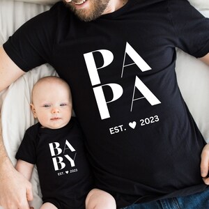 Op de afbeelding: Een zwart T-shirt met het woord "PAPA" in wit, verticaal gestapeld, en de tekst "EST. 2023" eronder. Een baby draagt een zwarte romper met het woord "BABY" in wit, verticaal gestapeld, en de tekst "EST. 2023" eronder.
