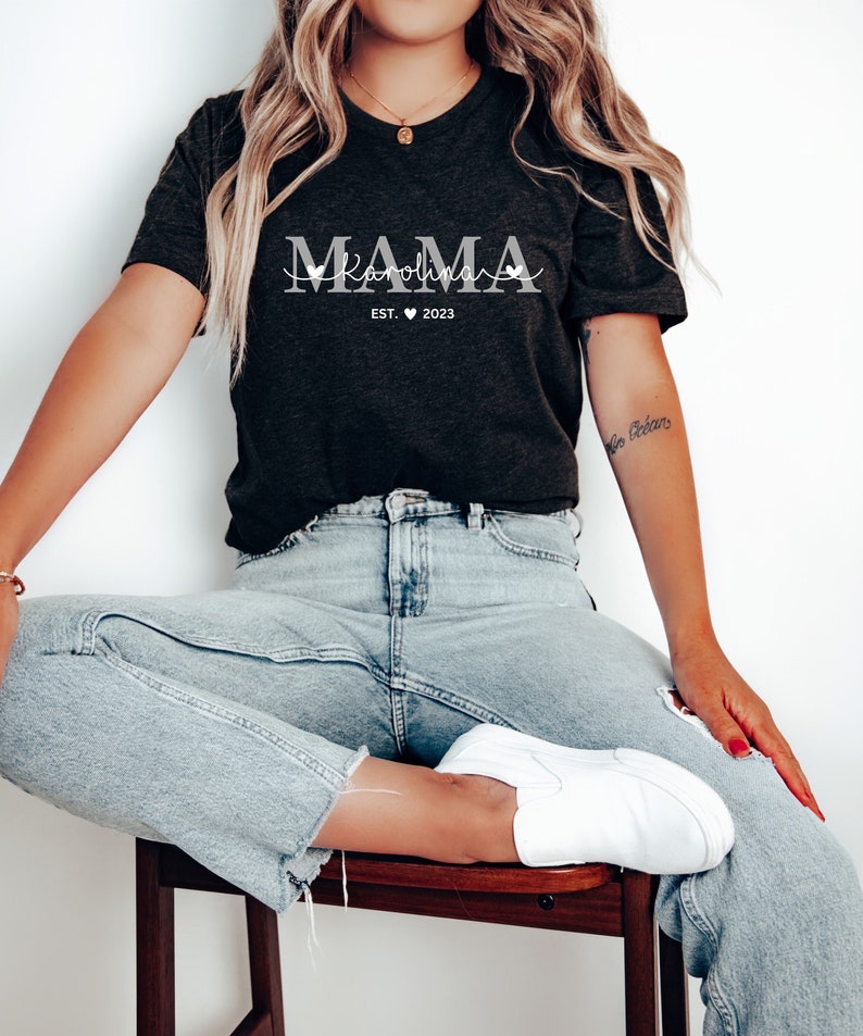 May include: A black t-shirt with the text "MAMA" in white with a script font underneath that says "Karolina EST. 2023".