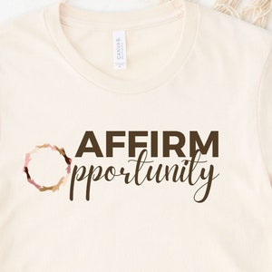 May include: A beige t-shirt with the text "AFFIRM opportunity" printed in brown. The text is surrounded by a circle of brown, pink, and gold.