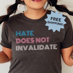 May include: A dark gray t-shirt with the text "Hate Does Not Invalidate" in blue and pink lettering. The text is in a bold, geometric font.