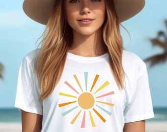 May include: A white t-shirt featuring a colorful sunburst design with orange, yellow, blue, and pink rays. The shirt is worn by a person wearing a wide-brimmed beige hat, with a beach and palm trees in the background.