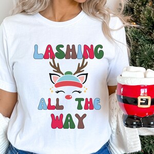 May include: White t-shirt with the text "LASHING ALL THE WAY" above a cartoon reindeer wearing a hat. The reindeer has eyelashes and a red nose. A red and black Santa mug with marshmallows is held in the hand.