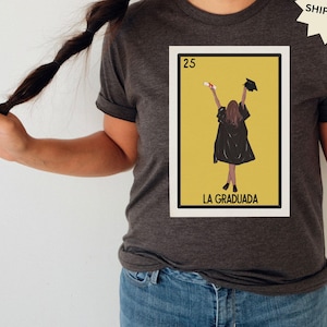 May include: A dark gray t-shirt with a graphic of a Loteria card featuring a woman in a graduation cap and gown. The card is labeled "25 La Graduada".