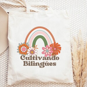 Op de afbeelding: Een witte tote bag met een regenboog en bloemen erop gedrukt. De tekst "Cultivando Biling&uuml;es" is onder de regenboog gedrukt.