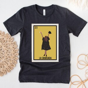 May include: A dark gray t-shirt with a yellow and white graphic of a person in a graduation cap and gown holding a diploma. The graphic is a parody of a Loteria card and says "La Graduada" at the bottom.