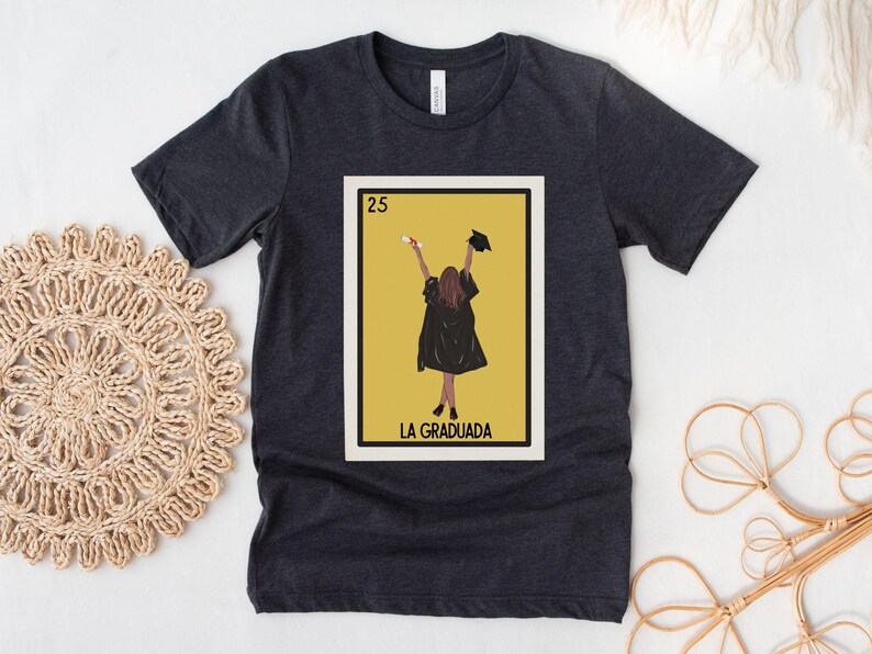May include: A dark gray t-shirt with a yellow and white graphic of a person in a graduation cap and gown holding a diploma. The graphic is a parody of a Loteria card and says "La Graduada" at the bottom.