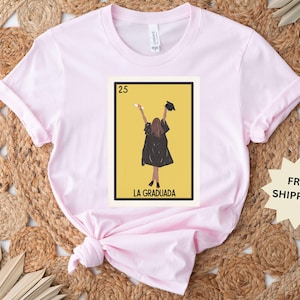 May include: Pink t-shirt with a graphic of a woman in a graduation cap and gown holding a diploma. The graphic is in the style of a Loteria card and says "La Graduada" and "25".