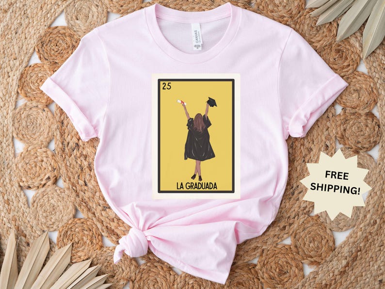 May include: Pink t-shirt with a graphic of a woman in a graduation cap and gown holding a diploma. The graphic is in the style of a Loteria card and says "La Graduada" and "25".