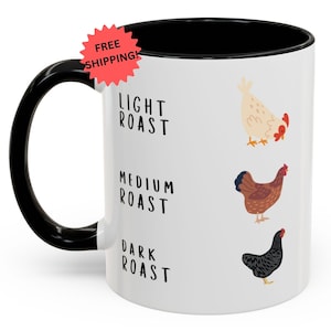 May include: White ceramic coffee mug with a black handle and interior. The mug features illustrations of chickens and the words "LIGHT ROAST", "MEDIUM ROAST", and "DARK ROAST". A red starburst sticker with the words "FREE SHIPPING!" is visible.