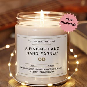 May include: A lit, cream-colored candle in a glass jar with a white label. The label reads "THE SWEET SMELL OF A FINISHED AND HARD-EARNED OD." The candle is 9 oz and has a 50-hour burn time. A pink starburst sticker says "FREE SHIPPING!"