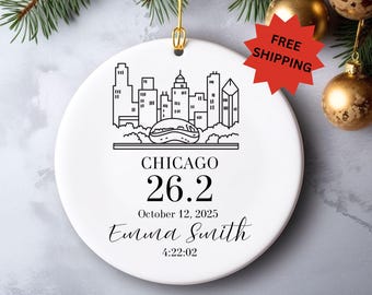 May include: White ceramic ornament with a black line drawing of the Chicago skyline, the text "CHICAGO 26.2", date, name, and time. A gold-colored loop is at the top for hanging. The ornament is decorated with a gold-colored ball and greenery.