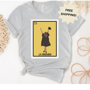 May include: A gray t-shirt with a graphic of a woman in a graduation cap and gown. The graphic is in the style of a traditional Mexican playing card. The text "25 LA GRADUADA" is printed below the graphic.