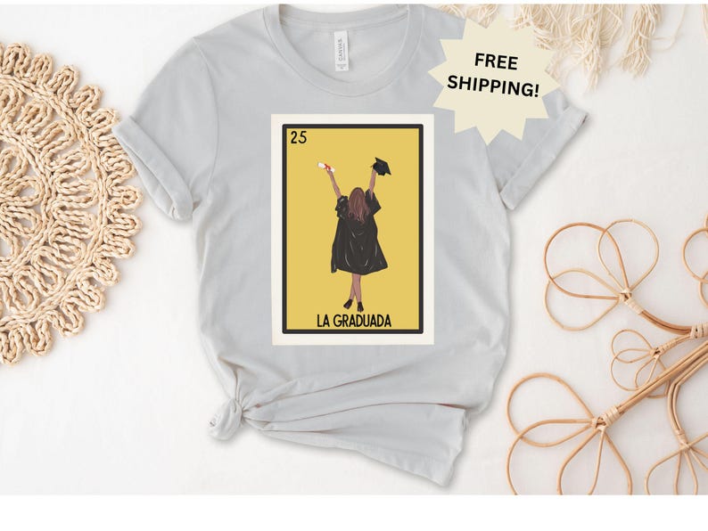 May include: A gray t-shirt with a graphic of a woman in a graduation cap and gown. The graphic is in the style of a traditional Mexican playing card. The text "25 LA GRADUADA" is printed below the graphic.