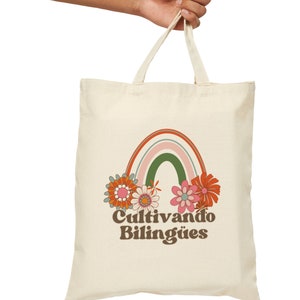 Op de afbeelding: Een witte canvas tote bag met een regenboog- en bloemenontwerp. De tekst "Cultivando Biling&uuml;es" is op de tas gedrukt.