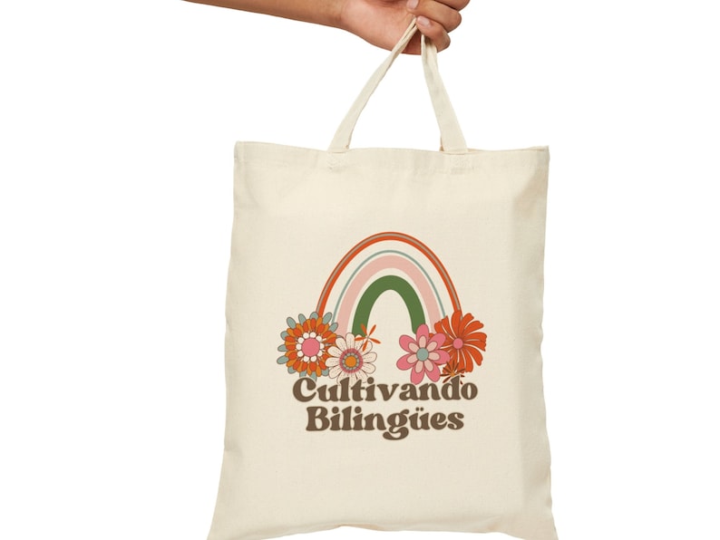 Op de afbeelding: Een witte canvas tote bag met een regenboog- en bloemenontwerp. De tekst "Cultivando Biling&uuml;es" is op de tas gedrukt.