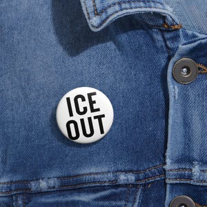 May include: A white button badge with the text "ICE OUT" in bold, black capital letters. The badge is pinned to a blue denim jacket. The badge is round and has a glossy finish.