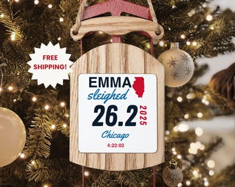May include: Wooden sled Christmas ornament with a red accent and a white rectangular insert. The insert reads "EMMA sleighed 26.2 Chicago 2025 4:22:02" with an Illinois state outline. The ornament hangs from a jute rope. A "FREE SHIPPING!" sticker is visible.