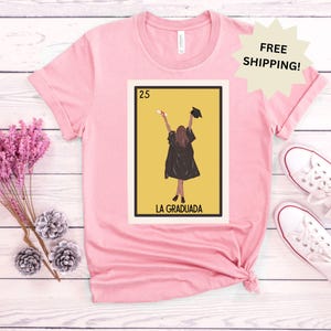 May include: Pink t-shirt with a graphic of a Loteria card featuring a woman in a graduation cap and gown. The card is labeled "La Graduada" and the number 25.