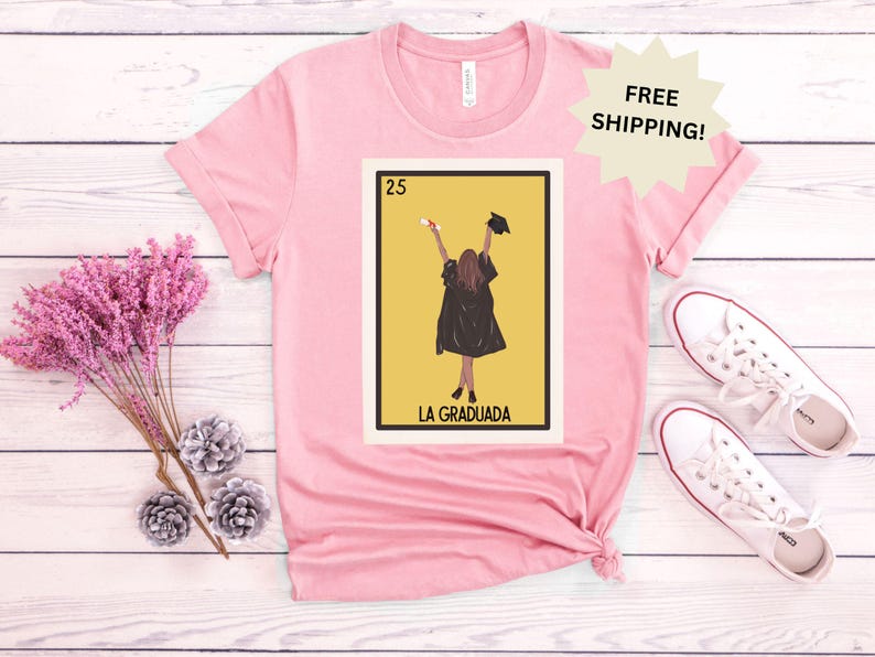 May include: Pink t-shirt with a graphic of a Loteria card featuring a woman in a graduation cap and gown. The card is labeled "La Graduada" and the number 25.
